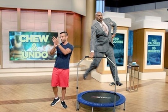 Steve Harvey bouncing on a rebounder trampoline on set while Jorge Cruise coaches him during the Chew & Undo segment